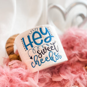 roll of toilet paper with hey sweet cheeks stitched on it in blue sitting on bed of pink feathers